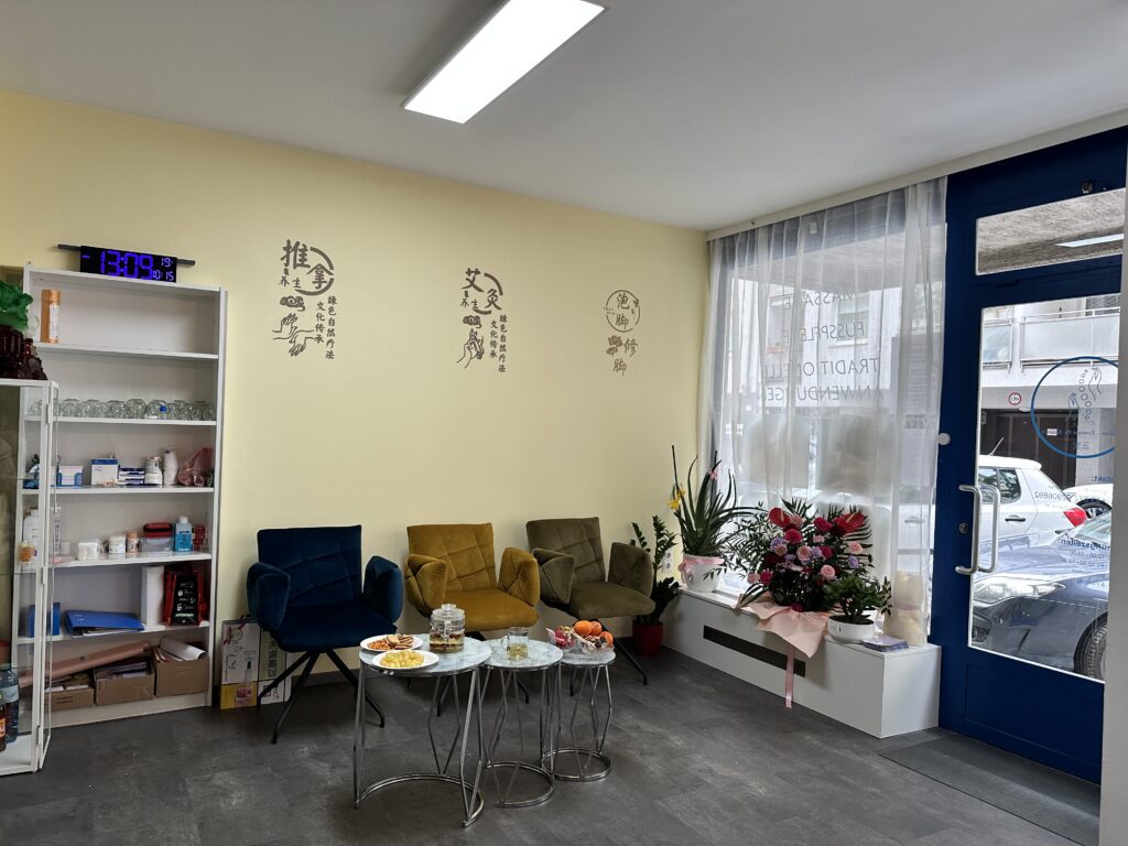 Wartebereich der Praxis Sanbao Balance mit Tee und traditionellen Elementen Heller Wartebereich der Praxis Sanbao Balance mit farbigen Sesseln, Teetisch, chinesischer Wandkunst und Blick auf die Straße durch ein Schaufenster